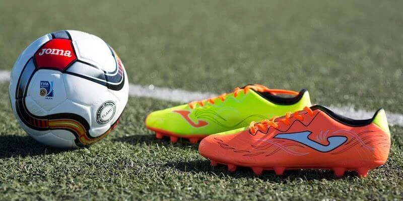 Sepatu bola Joma berwarna neon kuning dan oranye dengan bola Joma di lapangan rumput sintetis
